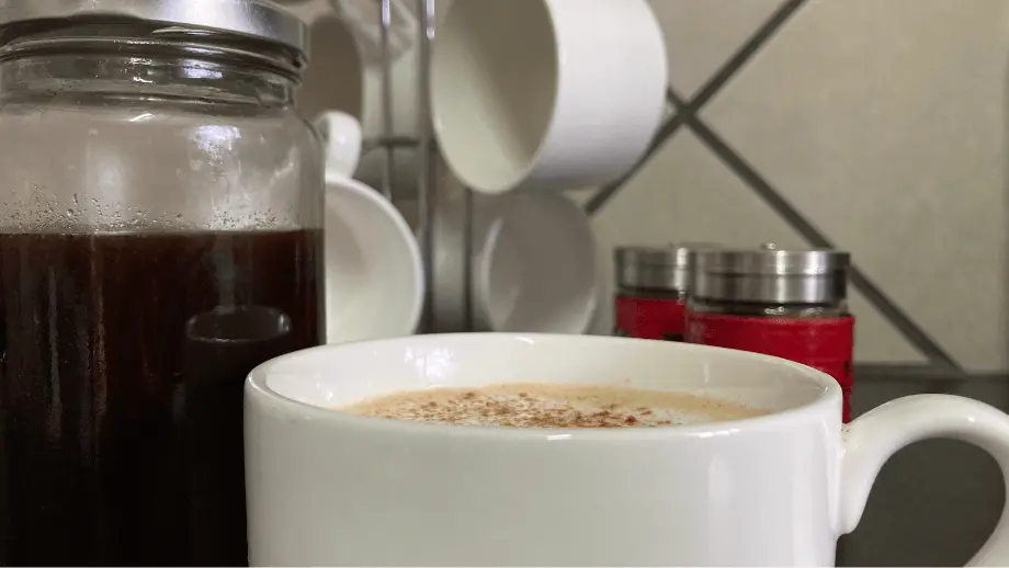 A pumpkin spice latte in a white mug front of a mason jar with pumpkin spice sauce.