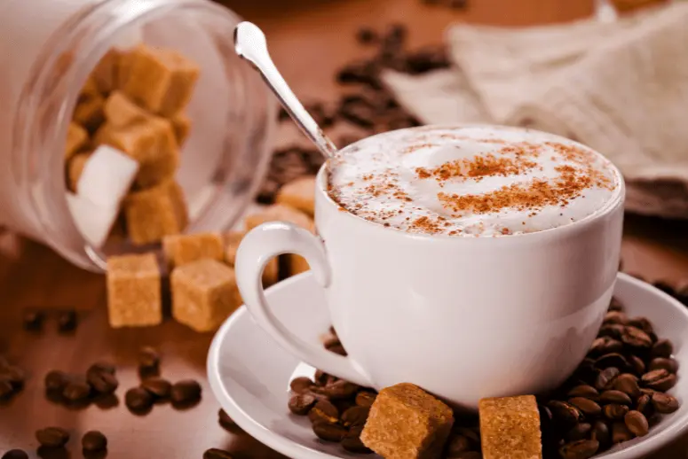 Cappuccino topped with cinnamon surrounded with coffee beans and sugar cubes.